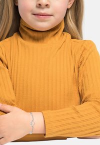 Maglione a collo alto giallo con coste, che mette in risalto un tessuto testurizzato. Braccialetto argento con un ciondolo a forma di cuore su una mano appoggiata sul braccio.