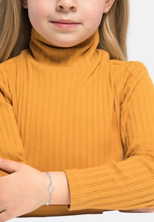 Maglione a collo alto giallo con coste, che mette in risalto un tessuto testurizzato. Braccialetto argento con un ciondolo a forma di cuore su una mano appoggiata sul braccio.