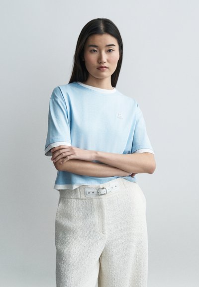 Femme aux longs cheveux foncés portant un chemisier bleu clair à manches courtes et un pantalon blanc texturé taille haute avec une ceinture, debout les bras croisés.