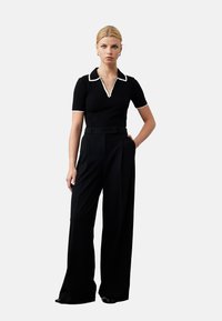 Zwarte polo met witte kraagdetails, gecombineerd met hoge taille zwarte brede broek. Model staat met één hand op de heup, op een neutrale achtergrond.