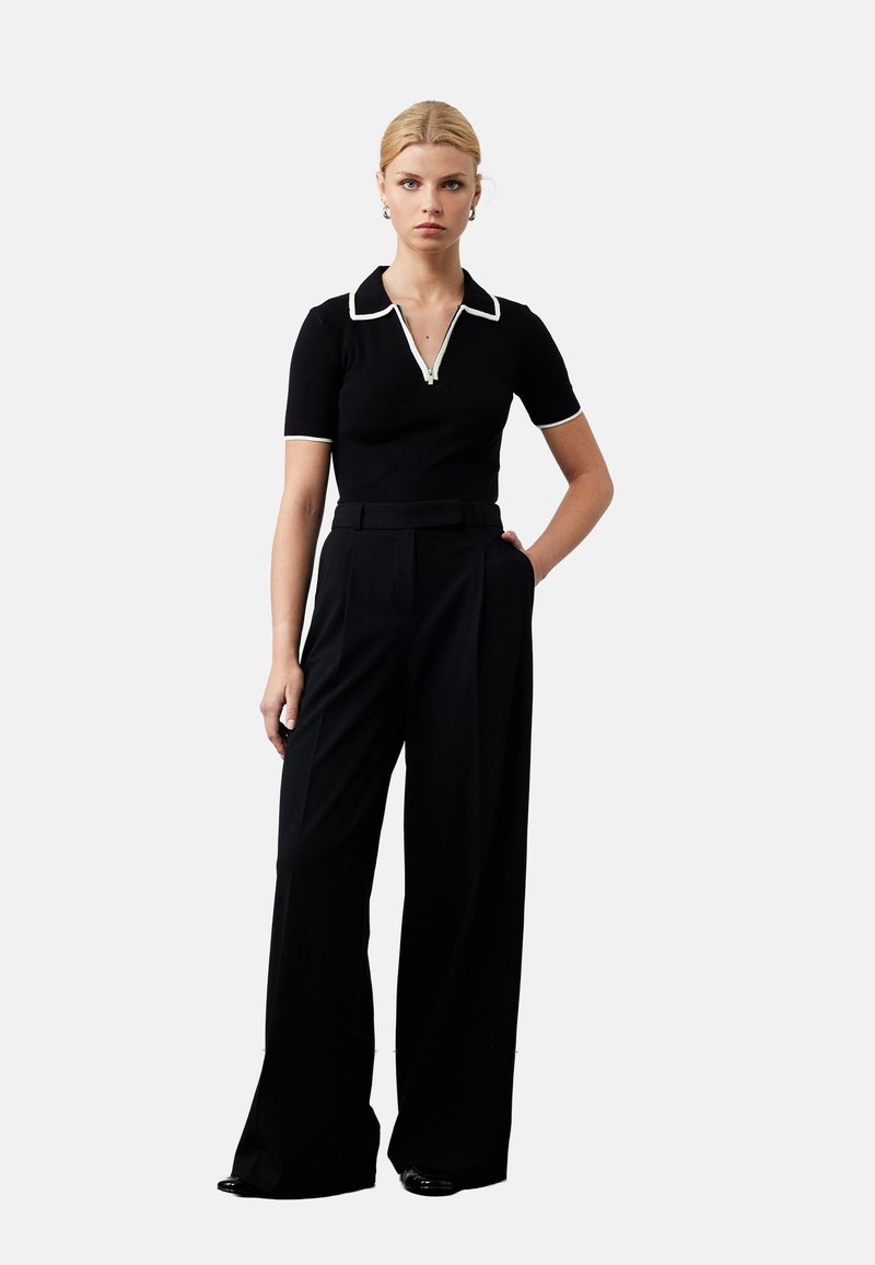 Zwarte polo met witte kraagdetails, gecombineerd met hoge taille zwarte brede broek. Model staat met één hand op de heup, op een neutrale achtergrond.