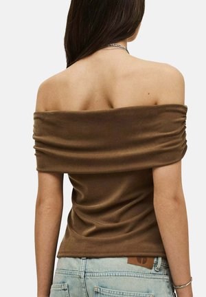 Donna con lunghi capelli scuri che indossa una maglietta marrone scollata sulle spalle e jeans azzurri chiari, vista da dietro su uno sfondo semplice.