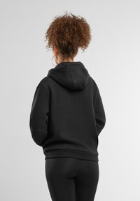 Felpa nera con tasca a marsupio, polsini e orlo a costine, dotata di cappuccio con cordino. Modello mostrato di spalle, abbinato a leggings neri.