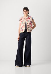 Blommönstrad väst i krämfärg med hög krage och framfickor, ihop med mörka vida jeans och ljusa skor.
