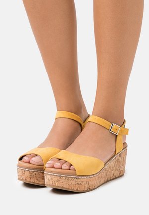 Keltaiset mokkanahkaiset platform-sandaalit avonaisella varvasmuotoilulla ja nilkkaremmillä. Korkkipohja, jossa on näkyvää tekstuuria ja ompelutietoja.