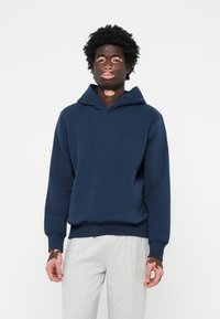 Felpa con cappuccio blu navy dal taglio morbido, con polsini a coste e un piccolo logo ricamato sul petto, abbinata a pantaloni felpati grigi chiari.
