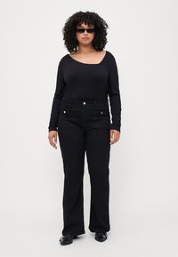 Black ribbed long-sleeve top with wide neckline, paired with high-waisted black flared pants featuring gold button accents. Black shoes and sunglasses.