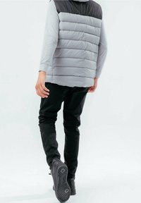 Vadderad väst i ljusgrått med en svart överdel, bärs över en grå långärmad tröja. Svarta byxor och mörka skor kompletterar outfiten.