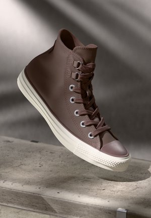 Hoge sneaker van bruin textuurleer met een witte rubberen zool. Heeft grijze veterogen en bijpassende veters, met minimale ontwerpelementen.