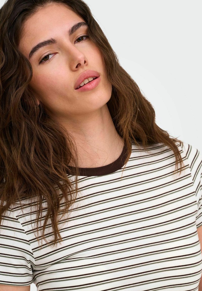 Jeune femme aux longs cheveux bruns ondulés portant un t-shirt blanc et noir à rayures, regardant légèrement vers le haut avec une expression neutre.
