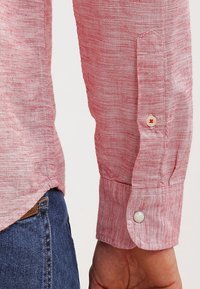 Camicia rossa con texture, dal taglio slim, con polsini a bottone e un delicato motivo intrecciato. Il tessuto appare leggero e traspirante.