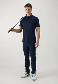 Camisola polo azul-marinho com colarinho e mangas curtas, combinada com calças azuis-marinho. O modelo segura um taco de golfe, vestindo sapatilhas brancas.