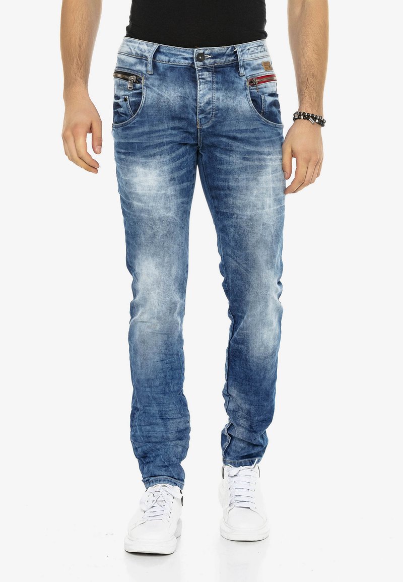 Denim spijkerbroek in een lichte blauwe wassing met versleten details, vijf zakken en ritsaccenten. Strak ontwerp met slanke pijpen, gedragen met witte sneakers.