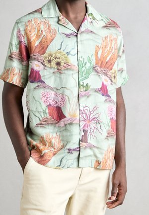 Mężczyzna noszący koszulę z krótkim rękawem i guzikami z nadrukiem pastelowej rafy koralowej i ukwiałów, w połączeniu z jasnymi spodniami, ręce spoczywają wzdłuż ciała.