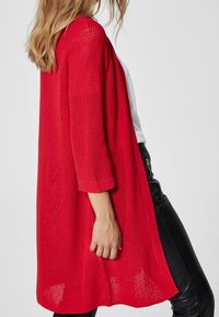 Röd stickad cardigan med trekvartsärmar, öppen front och en något längre bakdel, kombinerad med svarta slim-fit byxor.