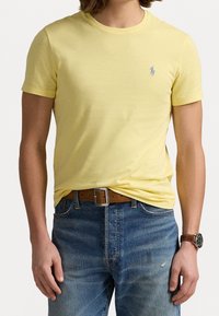 T-shirt gialla a maniche corte in cotone, con un piccolo logo blu sul petto. Indossata con jeans blu e una cintura marrone.