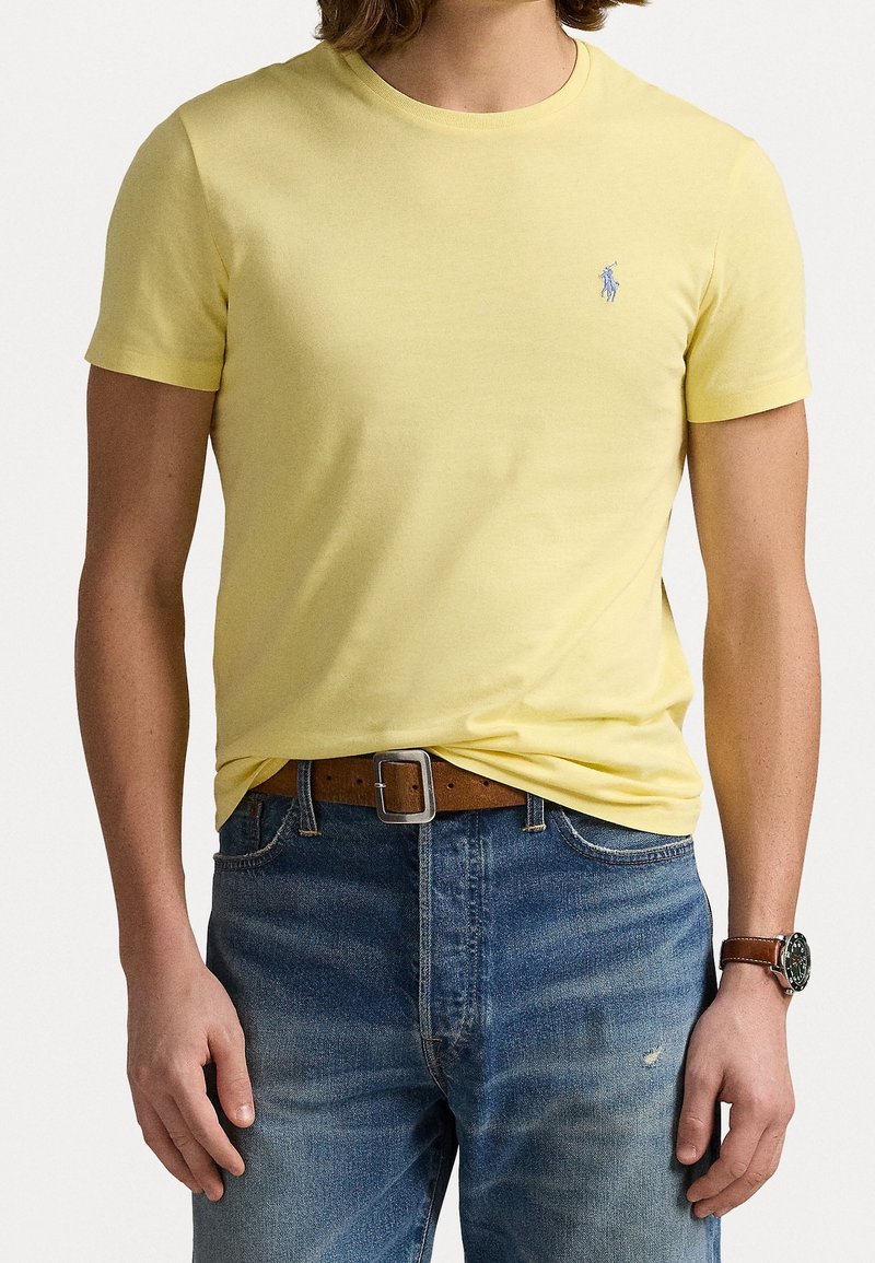 T-shirt gialla a maniche corte in cotone, con un piccolo logo blu sul petto. Indossata con jeans blu e una cintura marrone.