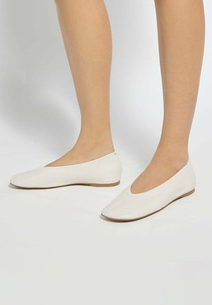 Voeten die eenvoudige witte leren ballerina's met lage hakken dragen, staand op een effen lichte achtergrond.