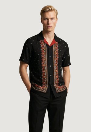 Uomo biondo che indossa una camicia nera a maniche corte con bottoni e motivi floreali verticali rossi e arancioni, pantaloni neri, in piedi con le mani nelle tasche.