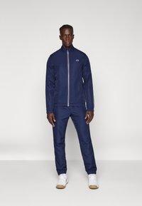 Mørkeblå træningsdragt i let materiale med en lynlåsjakke og elastiske manchetter; har et lille logo på brystet og matchende bukser.