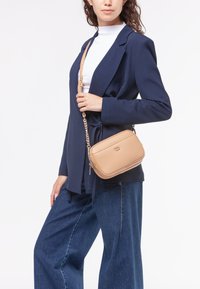 Borsa a tracolla in pelle beige con finitura testurizzata, forma rettangolare e tracolla in catena dorata, esposta accanto a un blazer scuro e denim.