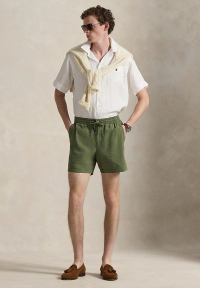 Ελαφρύ λευκό πουκάμισο από λινό, πράσινο υφασμάτινο μαγιό με υφή, κρεμ πλεκτό πουλόβερ περασμένο γύρω από τον λαιμό, καφέ σουέντ μοκασίνια και γυαλιά ηλίου.