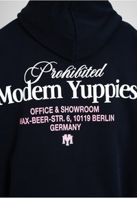 Sweat à capuche noir avec texte blanc et rose indiquant « Prohibited Modern Yuppies », adresse du bureau et du showroom à Berlin, et un petit logo stylisé en dessous.