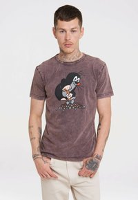 Man with tattoos wearing a faded purple t-shirt featuring a cartoon mole digging with a shovel, paired with light-colored pants.
