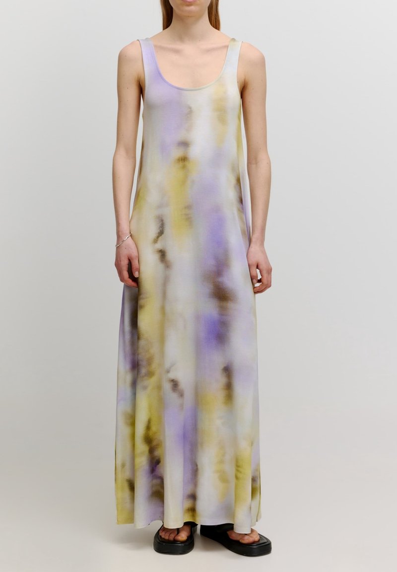 Femme portant une robe sans manches, longue jusqu'au sol, tie-dye aux couleurs pastel violet, jaune et blanc, avec des sandales à plateforme noires.
