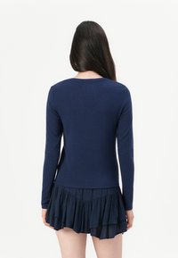 Kvinne med langt mørkt hår som vender bort, iført en marineblå langermet topp og et kort marineblått lagdelt skjørt mot en hvit bakgrunn.