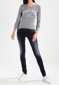 Top gris de manga larga con estampado en blanco "HILFIGER", combinado con jeans ajustados azul oscuro y zapatillas blancas; conjunto casual con una silueta entallada.