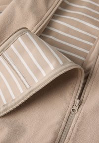 Beige fleece kledingstuk met ritssluiting, featuring een gestreepte binnenvoering met afwisselend beige en witte strepen. Glad oppervlak en zacht materiaal.