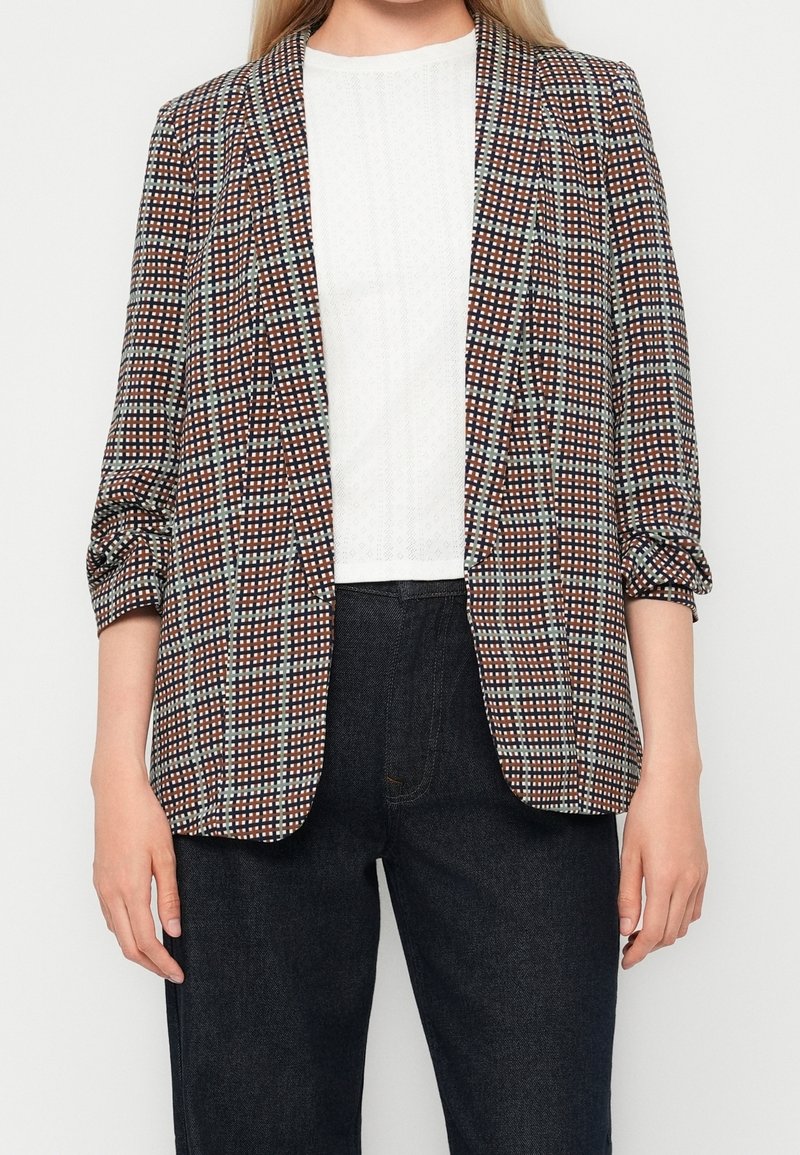 Femme portant un blazer à carreaux marron, blanc et noir avec des manches froncées sur un top blanc et un pantalon noir.