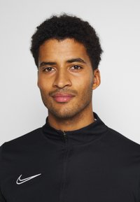 Zwarte sportjack met ritssluiting en hoge kraag, met een wit Nike-logo aan de linkerkant. Korte krullende haren en een neutrale achtergrond.