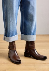 Une paire de bottines brunes avec boucle et fermeture éclair, portées avec un jean bleu retroussé sur un sol en bois clair.