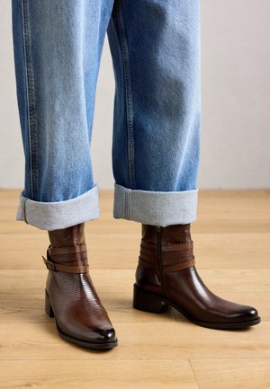 O pereche de ghete maro până la gleznă, cu cataramă și fermoar, purtate cu blugi albaștri răsuciți, pe un podea din lemn deschis.