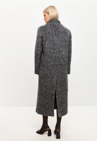 Abrigo largo gris con textura peluda, que presenta un collar alto y una abertura en la parte posterior. Zapatillas negras brillantes con tacón cuadrado son parcialmente visibles.
