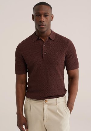 Homme portant un polo marron texturé à manches courtes et ajusté, ainsi qu'un pantalon beige, les mains dans les poches, faisant face à l'avant sur un fond uni.