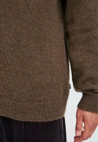 Brown knitted sweater with a textured finish, featuring ribbed cuffs and hem. The fabric appears cozy and thick. Small tag is visible.