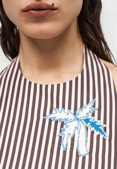 Κοντινό πλάνο στο κάτω μέρος του προσώπου και τον λαιμό μιας γυναίκας που φοράει ένα ριγέ τοπ με δέσιμο στον λαιμό και κεντημένο σχέδιο φοίνικα σε μπλε και άσπρο.