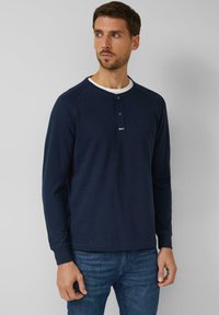 Mann trägt ein marineblaues Henley-Shirt mit langen Ärmeln und Knopfleiste, ein weißes Unterhemd und blaue Jeans, steht vor einer grauen Wand und blickt nach links.
