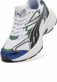 Sneaker bianco in rete con accenti verdi, blu e neri, dotato di sovrapposizioni in argento metallico, chiusura con lacci e dettagli testurizzati sulla suola.