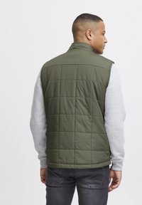 Olivgrüner, gesteppter Weste mit hohem Kragen, kombiniert mit einem grauen Langarmshirt. Verfügt über eine glatte Textur und minimalistisches Design.
