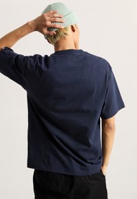 Camiseta de algodón azul marino con mangas cortas, cuello redondo y dobladillo recto. Modelo con un gorro de punto verde claro, visto desde atrás.