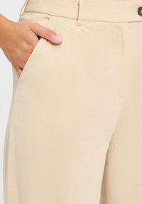 Beige op maat gemaakte broeken met een gladde textuur, voorzien van een zijzak en subtiele plooien. De tailleband heeft een lus voor een riem en een knoopsluiting.