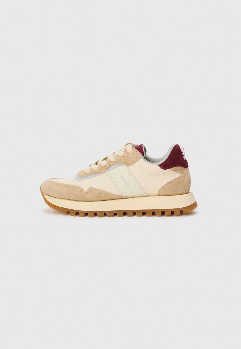 Beige und cremefarbene Sneaker mit beigen Wildlederakzenten, maroonfarbener Fersenschlaufe, weißen Schnürsenkeln und strukturiertem Gummisohle, Seitenansicht auf weißem Hintergrund.