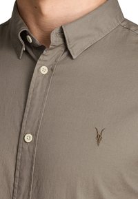 Beżowa koszula zapinana na guziki z kołnierzykiem i małym haftowanym logo czaszki barana na piersi.