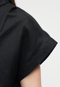 Κοντινό πλάνο του άνω βραχίονα ενός ατόμου που φοράει μαύρο μπλουζάκι με κοντό μανίκι και διπλωμένη μανσέτα, με φόντο ένα απλό φωτεινό υπόβαθρο.