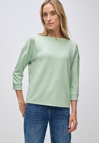Lys grønn, langærmet top med bådhals og ribbede detaljer. Blød tekstur og afslappet pasform, parret med blå jeans.