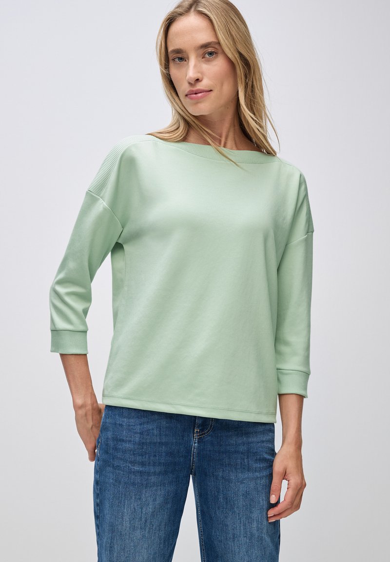Lys grønn, langærmet top med bådhals og ribbede detaljer. Blød tekstur og afslappet pasform, parret med blå jeans.